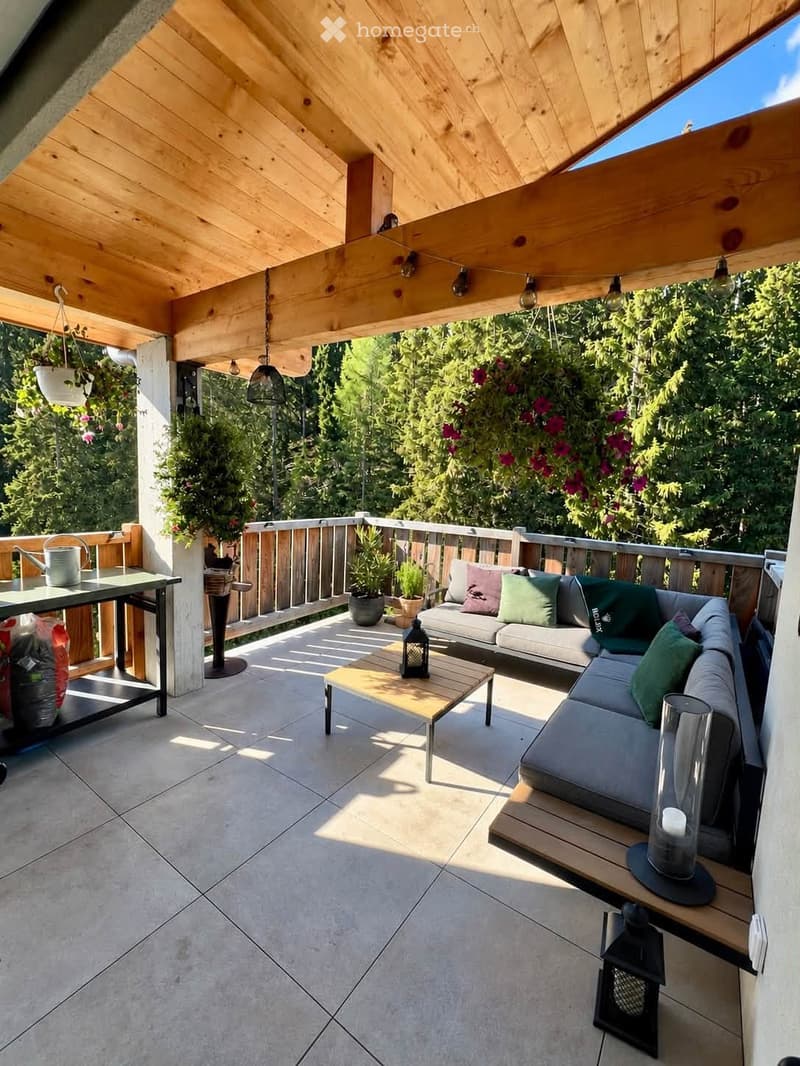 A modern wooden-covered balcony with a grey sectional sofa, coffee table, small table, potted plants, and decorative lanterns.
