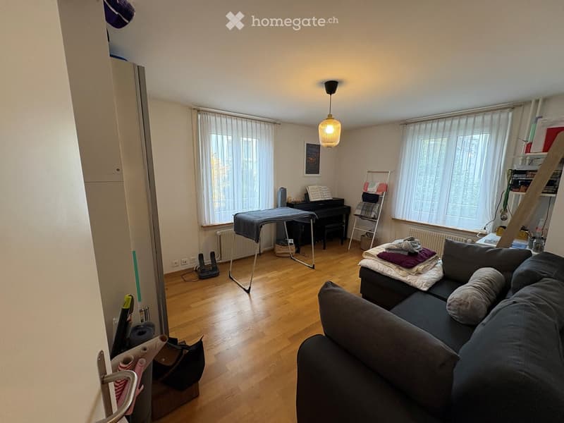 A well-lit living room with wooden flooring, a dark grey sofa, a piano, an ironing board, and a shelf with folded clothes. The room features large windows with white curtains and a hanging lamp.