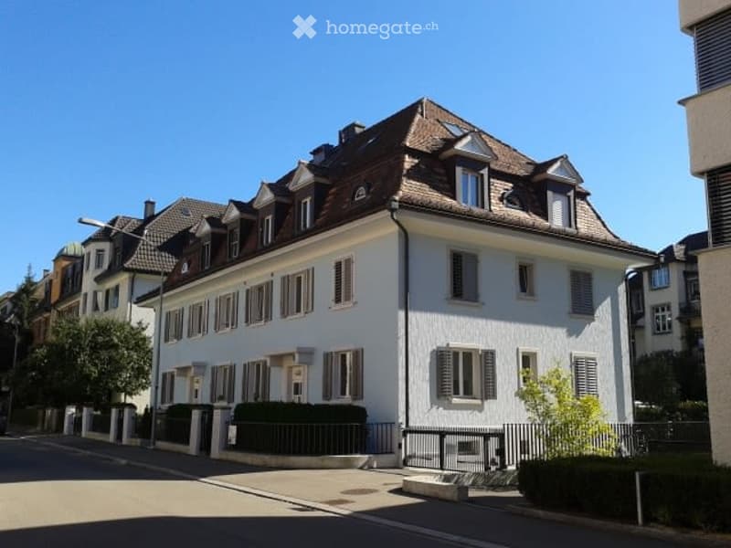 A multi-story residential building with a classic European architectural style, featuring white walls, dark brown roof, multiple windows with shutters, and a small garden area in front.