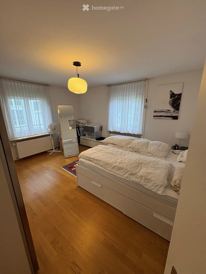 Cozy bedroom with wooden flooring, a double bed with white bedding, a mirror, a fan, and a table lamp.