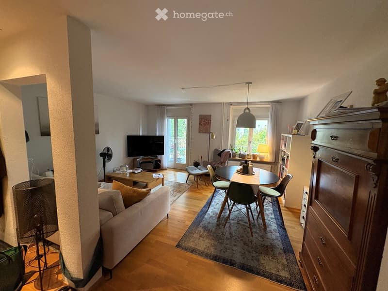 A well-lit living and dining area with wooden flooring, a sofa, coffee table, dining table with chairs, television, fan, and decorative items.