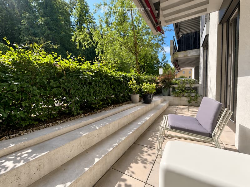 Balcony with a lounge chair, small table, and potted plants, surrounded by greenery and a concrete planter.