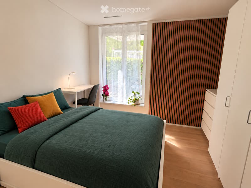 Modern bedroom with a green duvet, colorful pillows, a desk with a chair, a lamp, and a potted plant on the windowsill. The room features wooden flooring and white walls.
