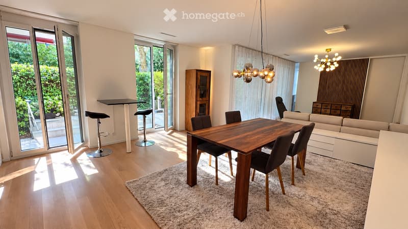 Modern living and dining area with a white sofa, wooden dining table, chairs, bar stools, and a chandelier.