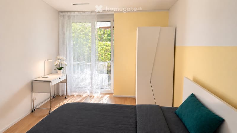 A modern, furnished room with a sofa, bed, small table, and a lamp. The room features a large window with sheer curtains, a view of greenery, and a colorful wall design.