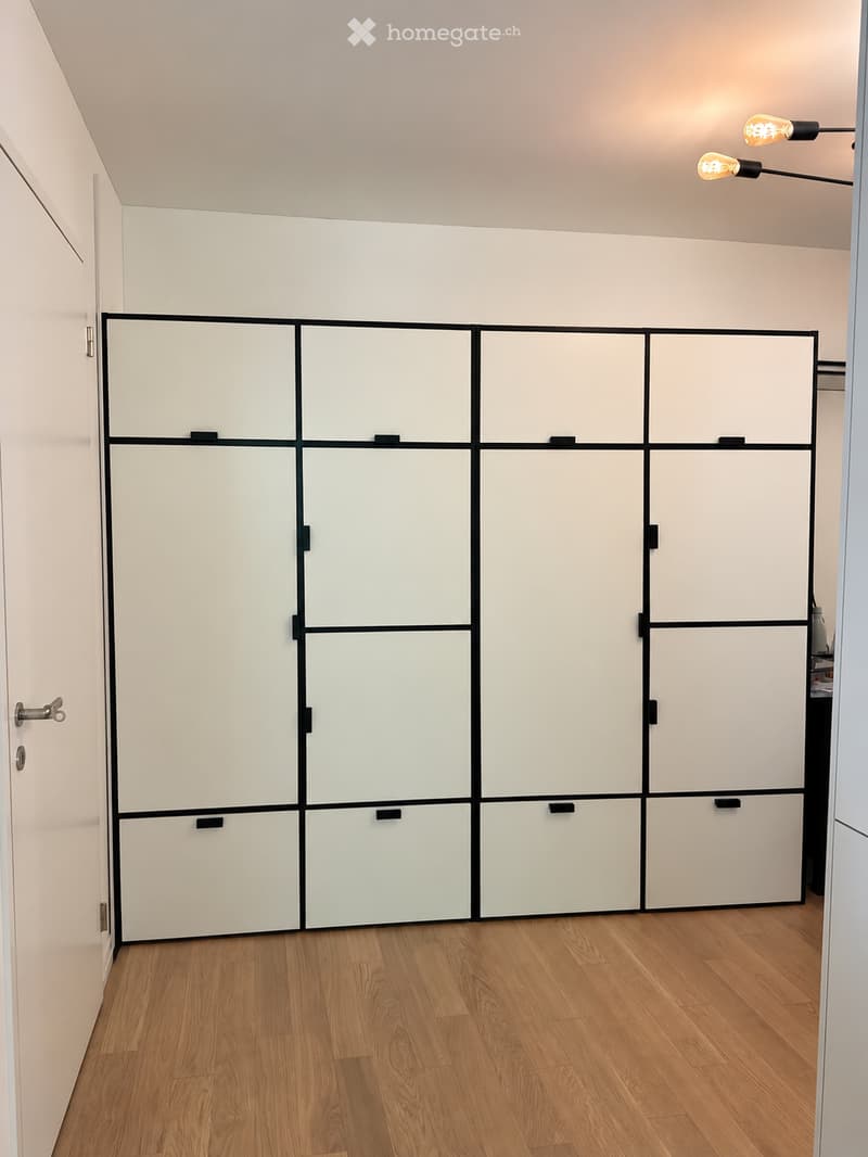 Empty room with light-colored walls, wooden flooring, and a large white cabinet with black trim. The room has a modern, minimalist design with no visible furniture or personal items.