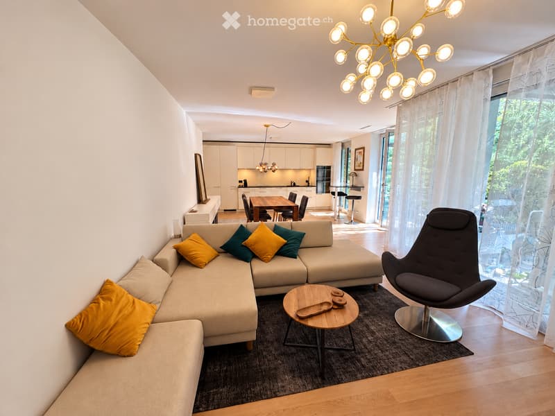 Modern living room with a beige sectional sofa, colorful throw pillows, a wooden coffee table, a sleek black chair, and a dining area with a wooden table and chairs. The space features large windows with sheer curtains, a chandelier, and a view of greener