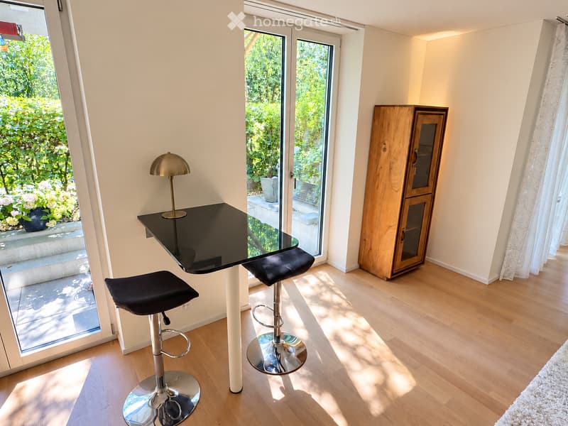 Bright room with wooden flooring, large windows, a black countertop with two black stools, a wooden cabinet, and a gold lamp on the table.