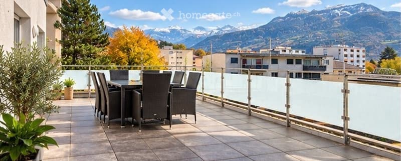Balcony with a modern glass railing, overlooking a scenic mountain view. A dining set with four chairs and a table is placed on the tiled floor, surrounded by potted plants.