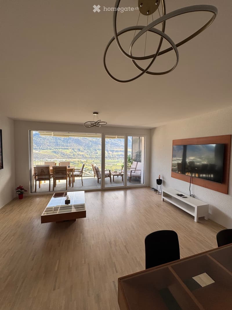 Modern living room with wooden flooring, large windows offering a scenic view, a coffee table, chairs, a dining table, and a TV screen on a wooden stand.