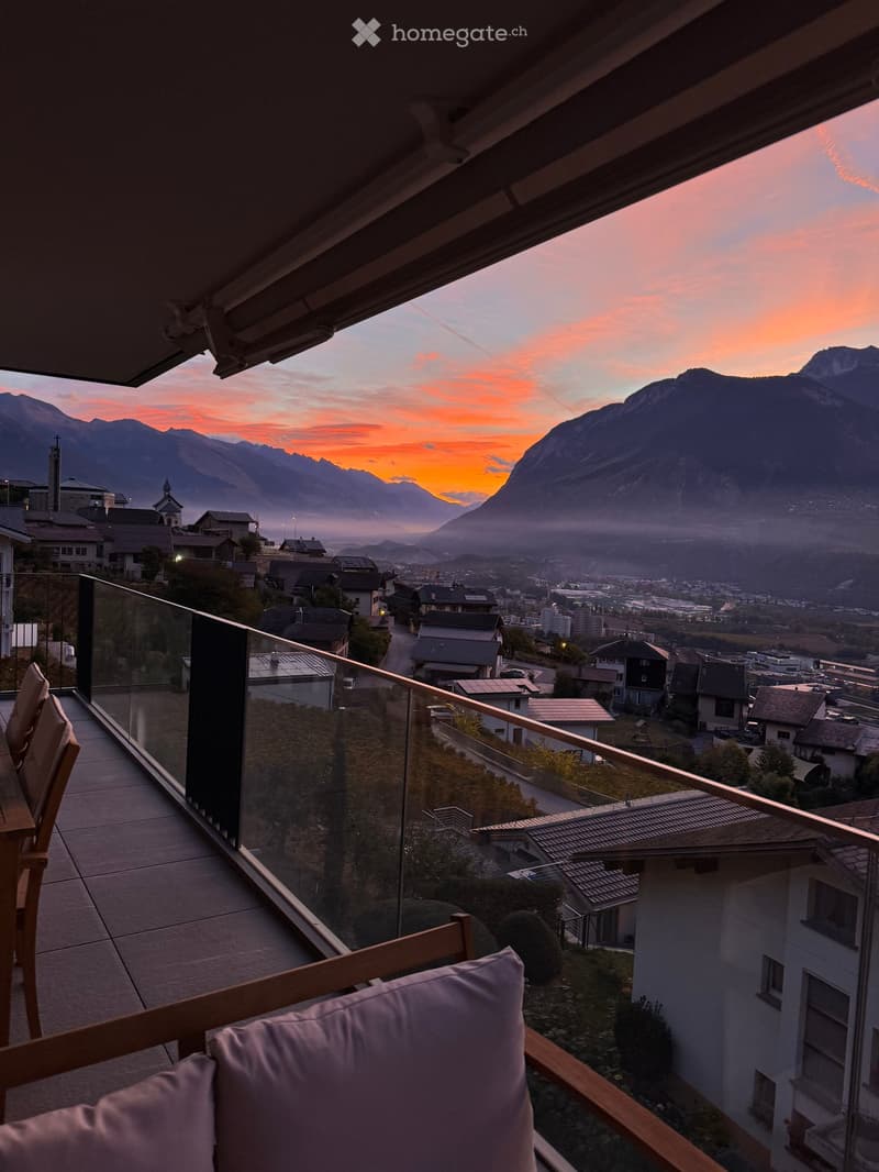 Balcony with glass railing, wooden chairs, and a table, overlooking a town and mountains at sunset.