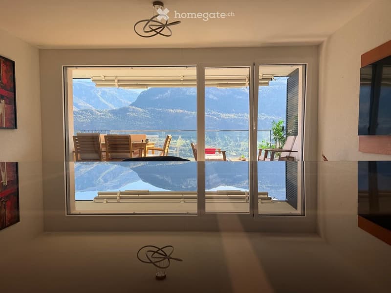 Modern room with large glass doors opening to a balcony, overlooking a scenic mountain view. Features include a sleek hanging light fixture, a small table with chairs, and a flat-screen TV on the wall.