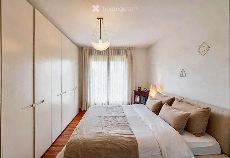 Modern bedroom with a large bed, nightstand, books, pendant light, and built-in wardrobe.
