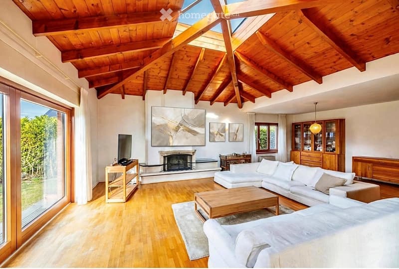 Bright living room with wooden floors, exposed wooden ceiling beams, large windows, white sectional sofa, coffee table, fireplace, TV, and wooden cabinetry.