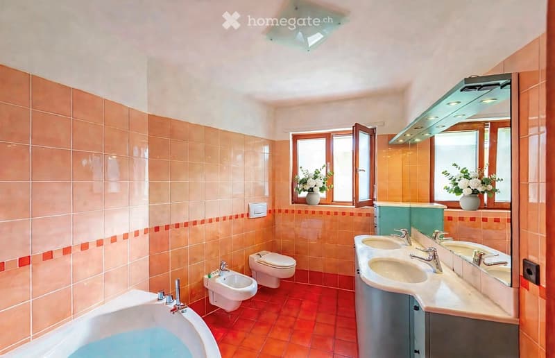 Bright bathroom with peach-colored tiles, a bathtub, bidet, toilet, double sink with mirrors, and vases on windowsills.
