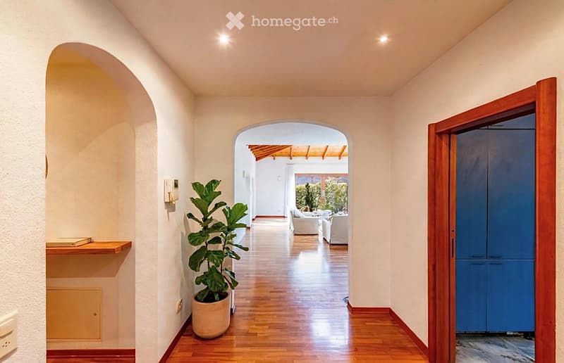 Elegant hallway with wooden flooring, arched doorways, and a potted plant, leading to a bright living area with white furniture and a view of greenery outside.