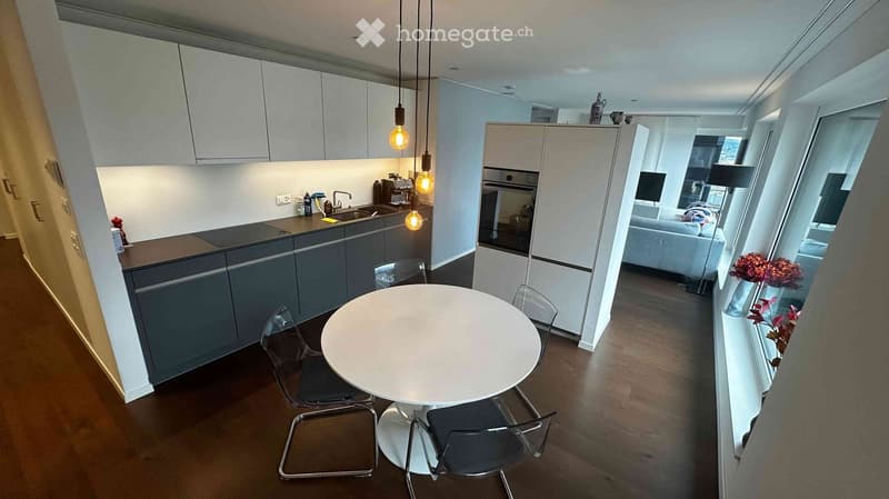 Modern kitchen with grey cabinets, white countertop, sink, and built-in oven. A round white dining table with four transparent chairs is placed in the center. A living area with a sofa and TV is visible through an open doorway.