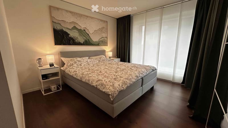 Modern bedroom with a large bed, nightstand, lamp, and a large abstract painting on the wall. The room features large windows with sheer curtains and a dark wooden floor.