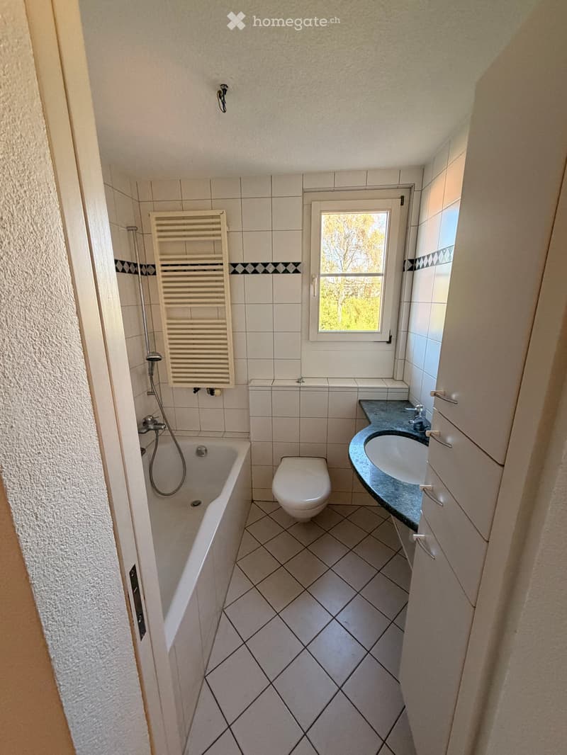 Bathroom with white tiled walls and floor, featuring a bathtub, toilet, sink, and radiator. A window provides natural light.