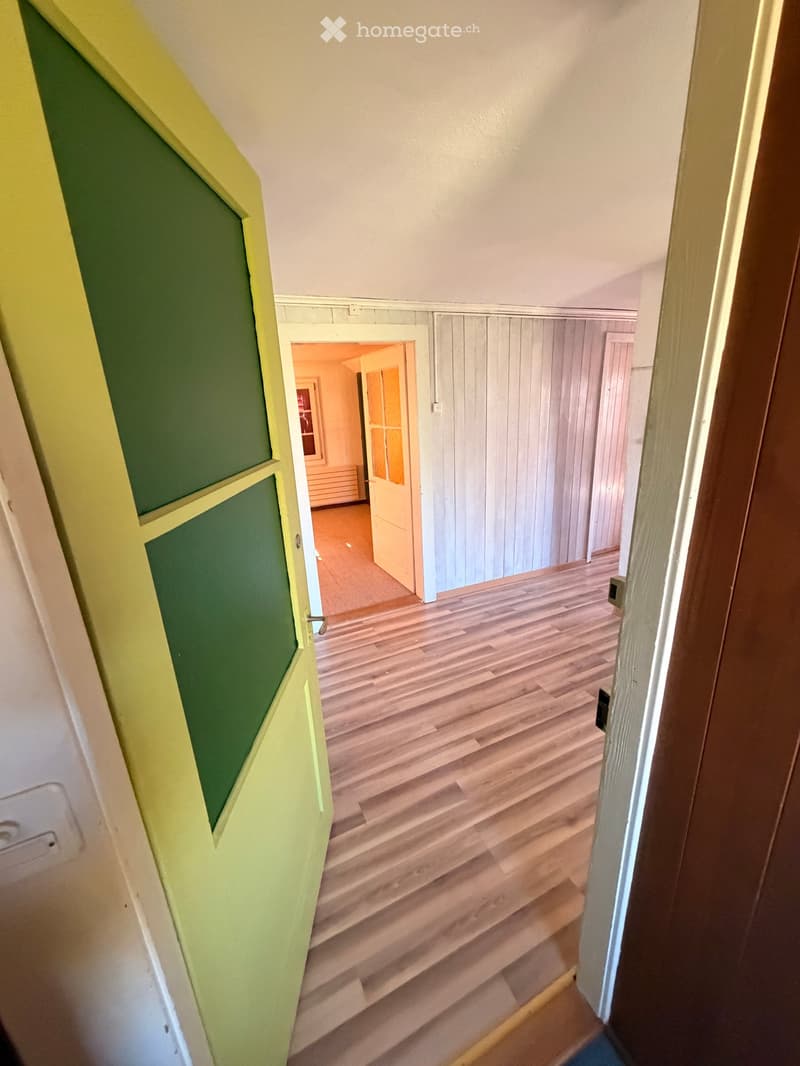 A narrow hallway with wooden flooring, white walls, and a green door on the left. An open doorway leads to another room with wooden paneling and a visible piece of furniture.
