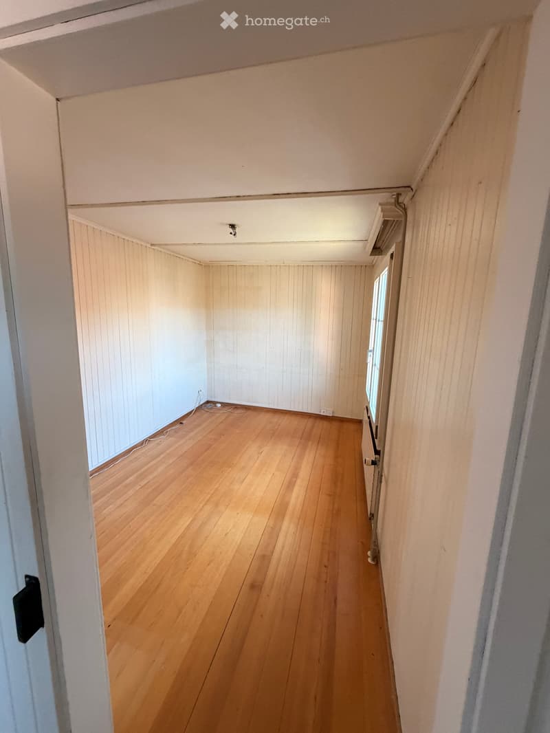 Empty room with wooden flooring, white walls, and a small window letting in natural light. The room has a simple, clean design with no furniture or decorations.