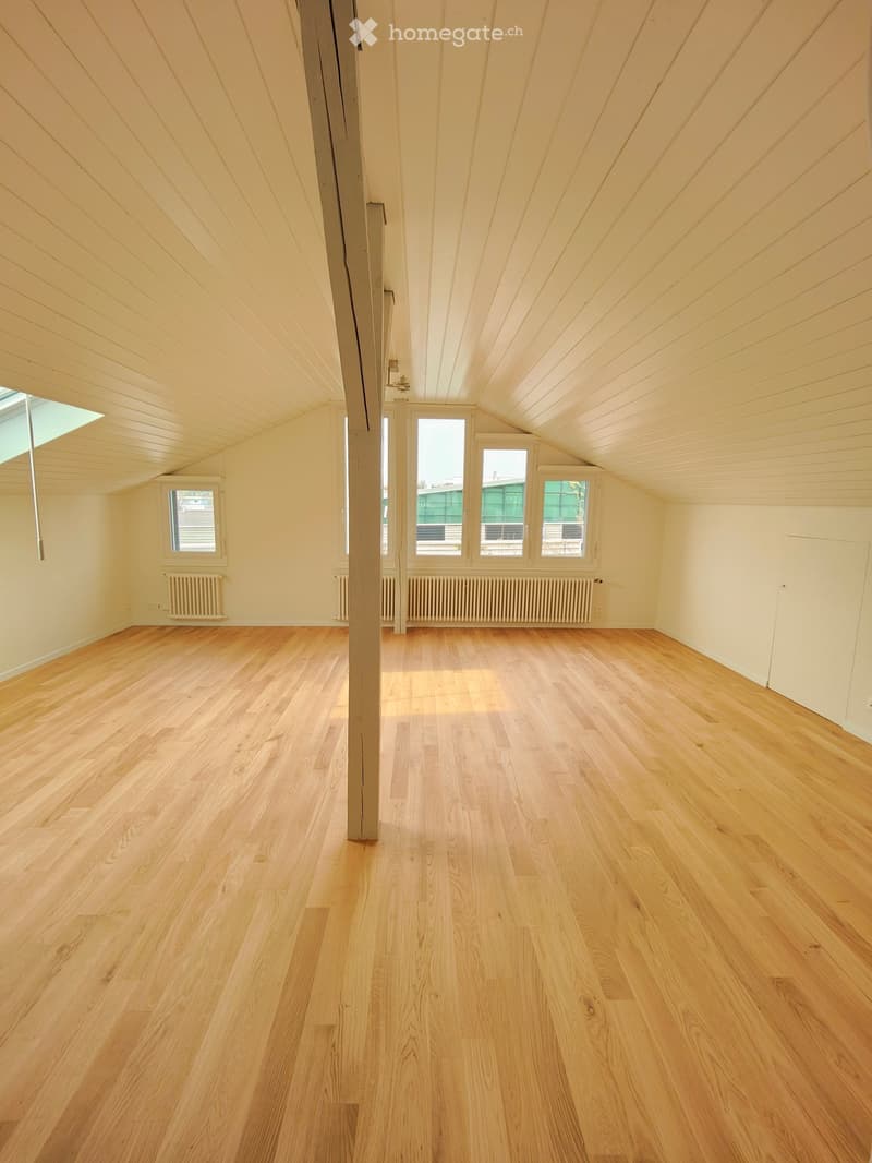 Empty room with wooden flooring, light-colored walls, and large windows allowing natural light. The ceiling has exposed wooden beams, and there is a central support column.