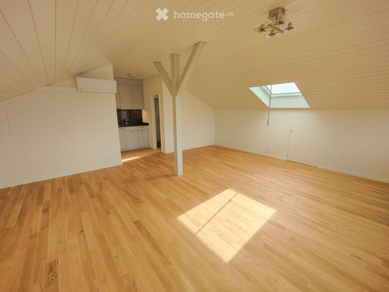 Empty room with wooden flooring, white walls, and a skylight window allowing natural light. The room features a slanted ceiling with exposed wooden beams and a small kitchen area visible in the background.