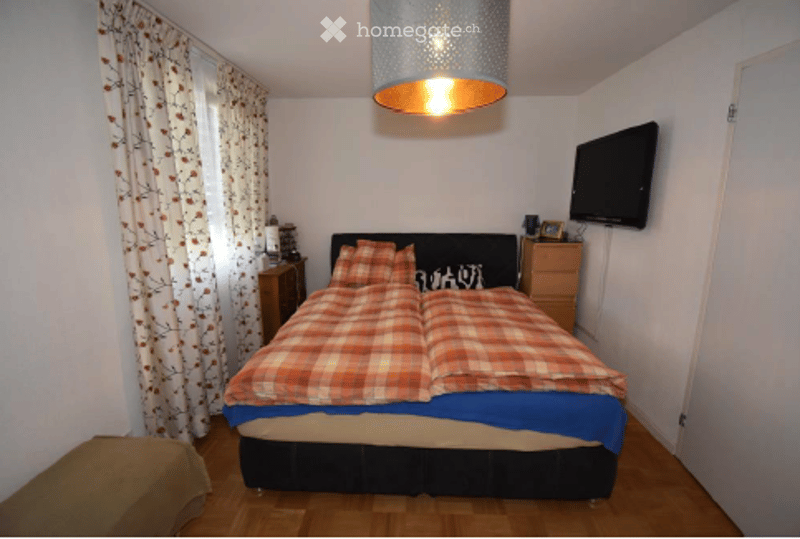 Cozy bedroom with a large bed, plaid bedspread, nightstands, a television mounted on the wall, and a hanging pendant light.