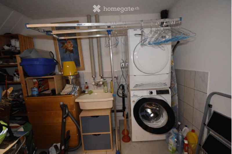 The image shows a small bathroom with a sink, washing machine, and various storage items. There is a wooden cabinet with drawers, a plastic hanger rack above, and cleaning supplies on the floor.
