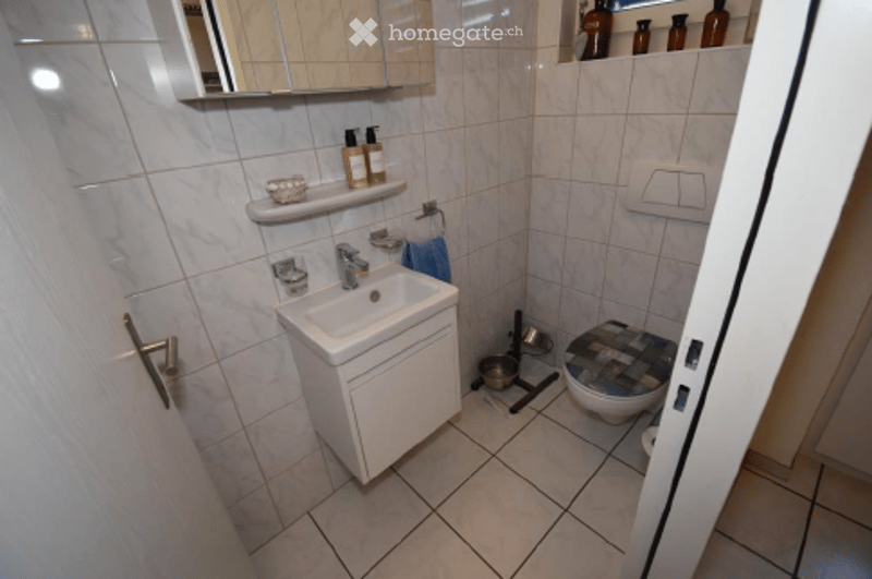 Modern bathroom with white tiles, a white sink, a mirror, a toilet with a patterned seat cover, and a towel hanging on a rack.