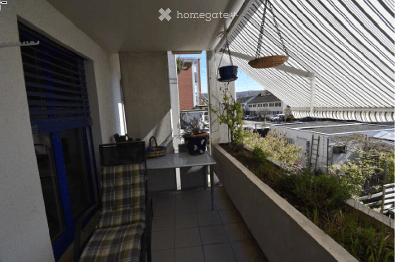 Balcony with a small table, chair, and potted plants, overlooking a street with buildings.