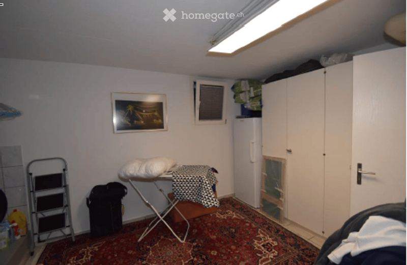 A small, furnished room with a carpeted floor, a white cabinet, an ironing board, a picture frame on the wall, and a trash bin. The room includes a folding chair and a stool.