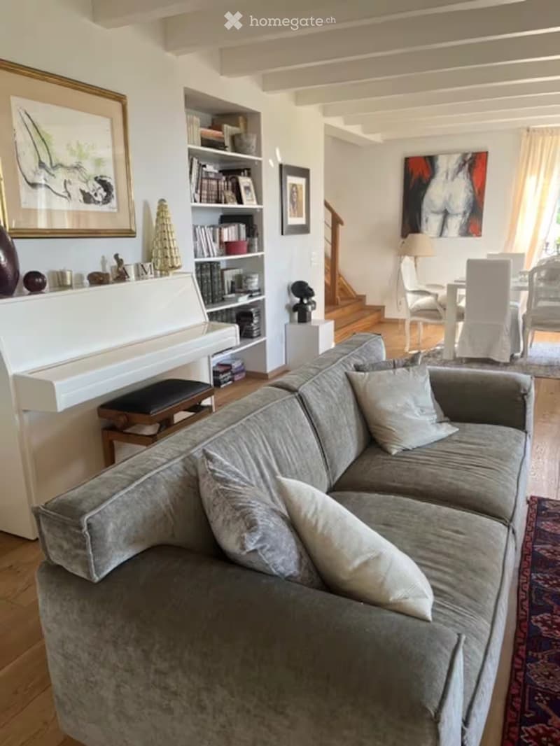 A cozy living room with a grey sectional sofa, decorative pillows, a bookshelf filled with books, a small table with a lamp, and a staircase leading to another level.