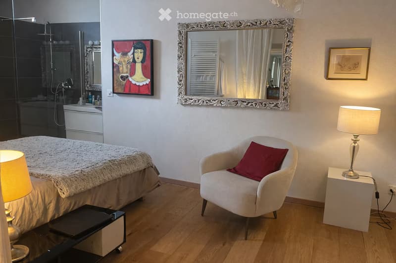 Cozy bedroom with a large bed, a white armchair with a red pillow, a table lamp, a nightstand, a large ornate mirror, and a framed picture on the wall. The room features wooden flooring and a modern, elegant design.