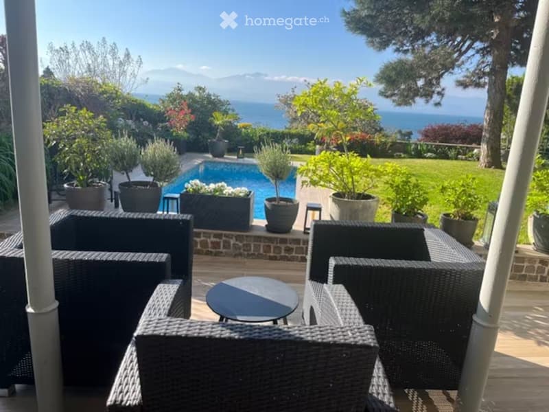 Outdoor patio with wicker chairs and a small table, overlooking a swimming pool and scenic ocean view with surrounding greenery and trees.
