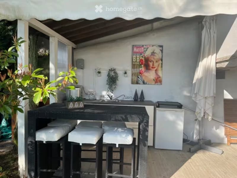 A covered outdoor terrace with a black table, three white-cushioned stools, a small refrigerator, potted plants, and a large umbrella.