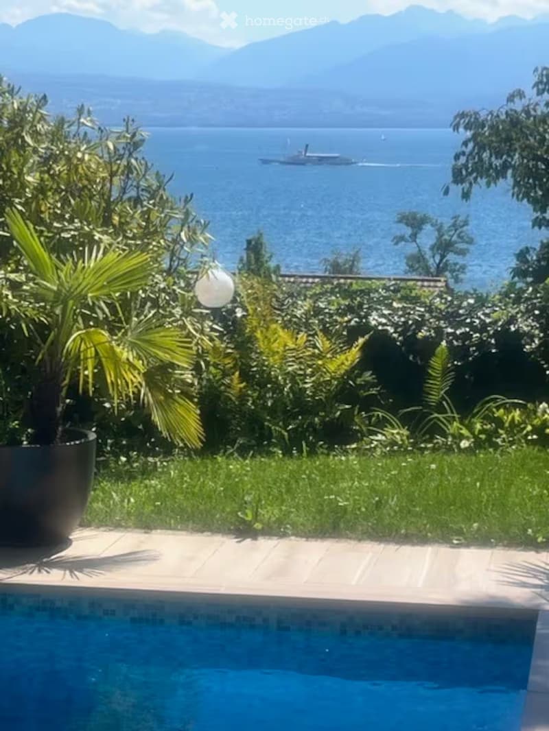 A serene outdoor area with a swimming pool, surrounded by lush greenery and a scenic view of a lake and mountains in the background.