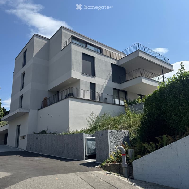 Modern two-story house with white facade, large windows, and multiple balconies, surrounded by greenery and a concrete driveway.