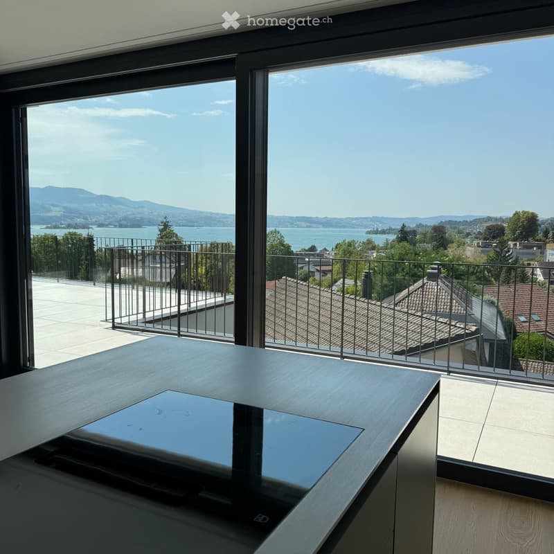 Modern room with large windows offering a scenic view of a lake and mountains, featuring a sleek wooden table with a laptop on it.