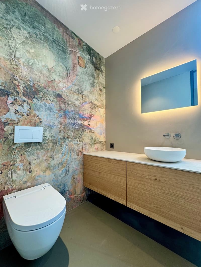 Modern bathroom with a white toilet, a wooden vanity with a white sink, a large mirror with built-in lighting, and a decorative textured wall.
