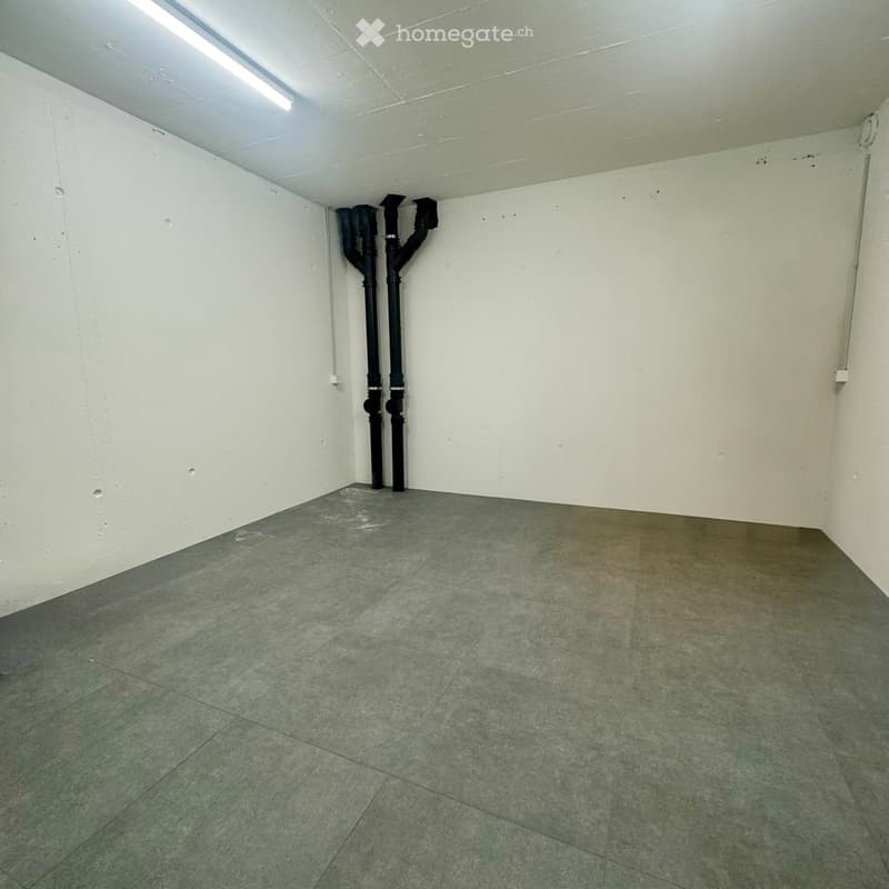 Empty room with concrete floor and white walls, featuring exposed black pipes and a single fluorescent light fixture on the ceiling.