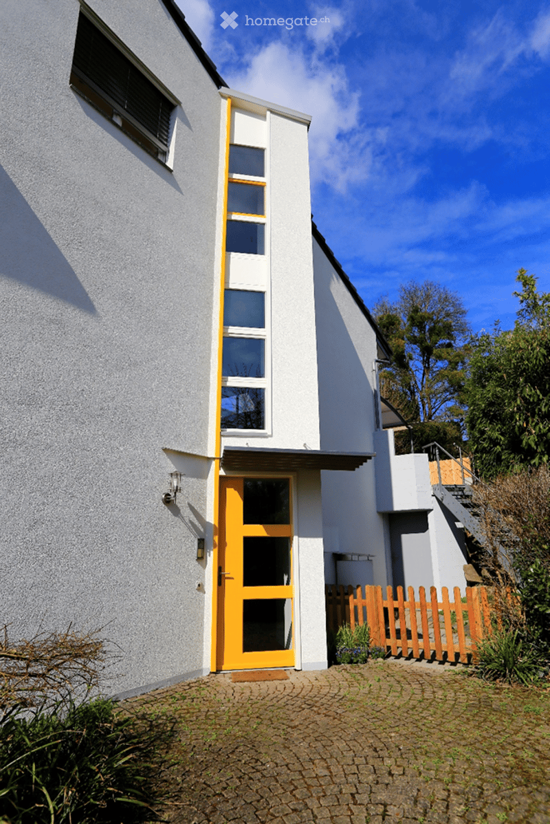 Modern two-story house with grey and white exterior, orange front door, and raised ground floor entrance.