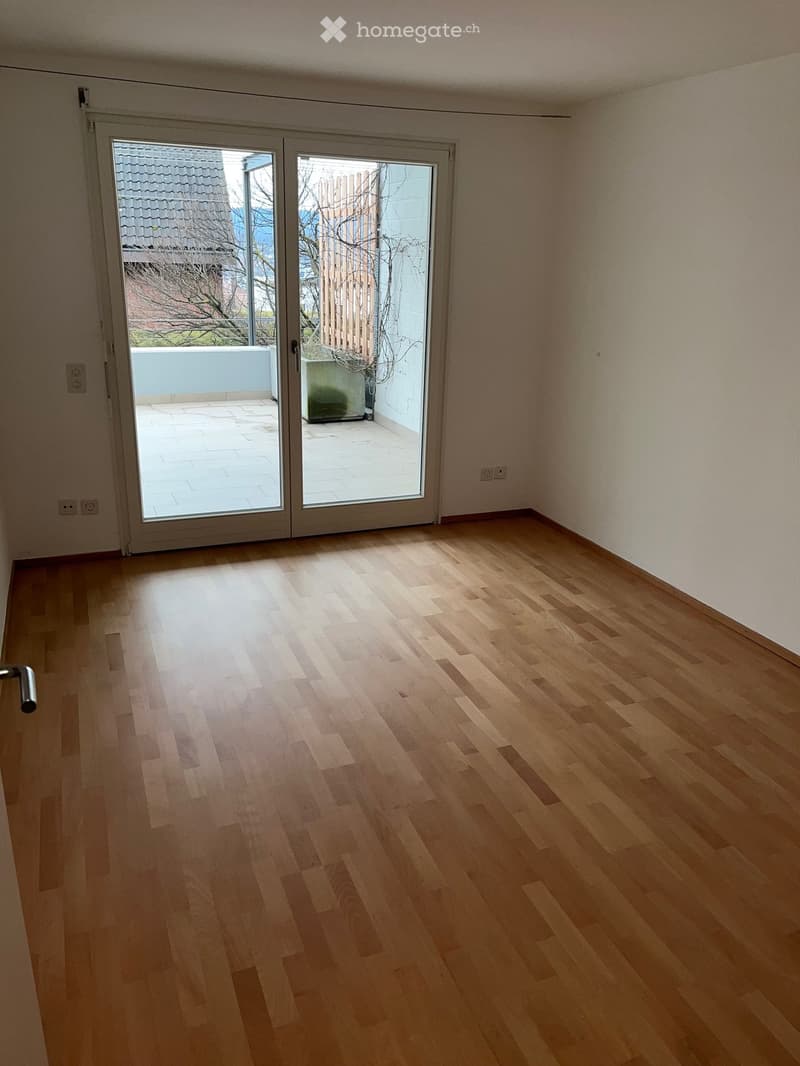 Empty room with light wooden flooring, white walls, and a large sliding glass door leading to a balcony or outdoor area.