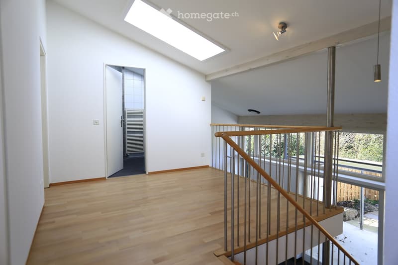 Bright, modern hallway with wooden flooring, white walls, and a staircase leading upwards. Features include a skylight, pendant lighting, and a view of the outdoor area through large windows.