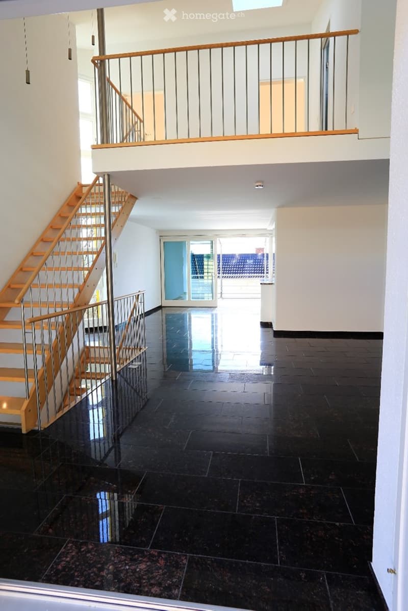 Open, modern hallway with polished black tiled flooring, wooden staircase with metal railing, and a balcony overlooking the space.