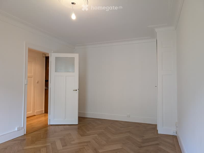 Empty room with white walls, wooden herringbone flooring, and a white door with frosted glass panel. The room has a ceiling light fixture and visible electrical outlets.