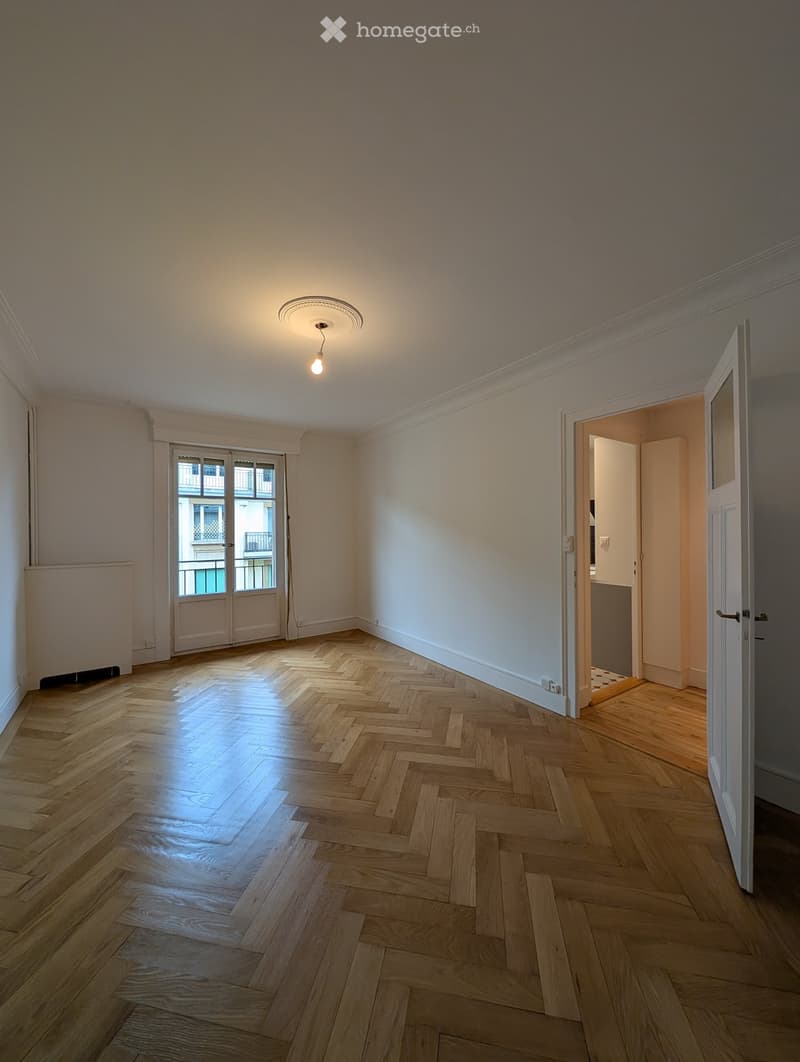 Empty room with wooden herringbone flooring, white walls, and a ceiling light fixture. Features a large window with a view of a balcony and an open doorway leading to another room.