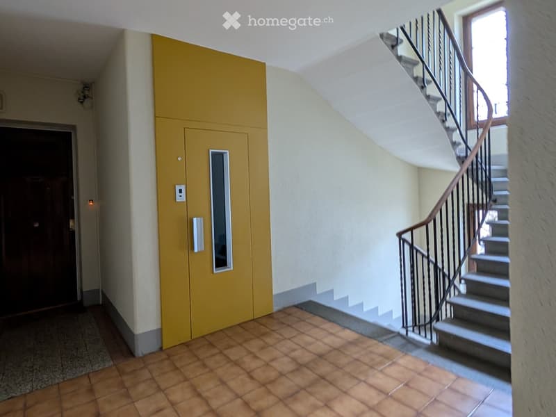 Hallway with a staircase leading upwards, featuring a yellow elevator door on the left and a wooden door on the far left. The floor is tiled with terracotta tiles, and the walls are painted in a light cream color.