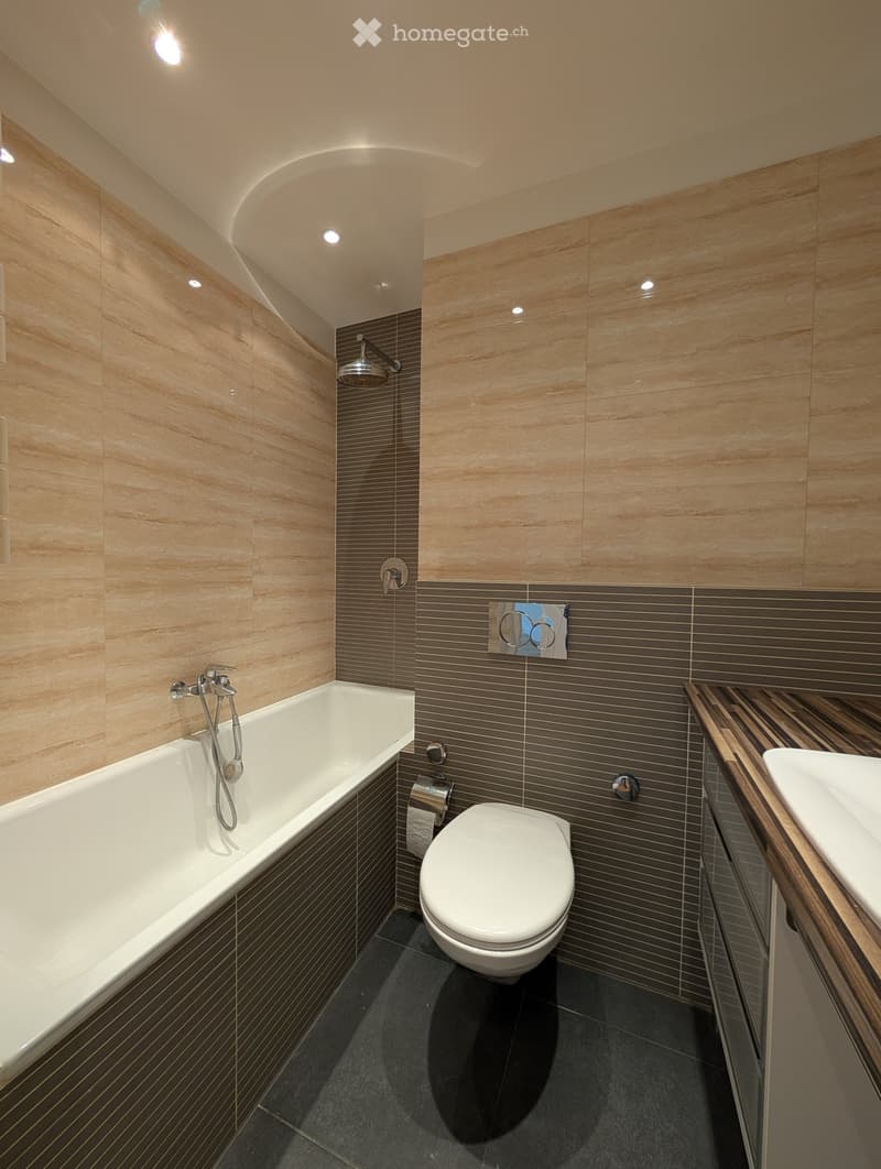 Modern bathroom with a bathtub, toilet, sink, and showerhead. Features light wood paneling and dark tiled accents.