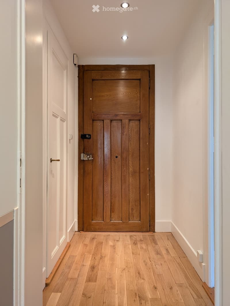 A narrow hallway with light wooden flooring, white walls, and a large wooden door at the end. The hallway has recessed ceiling lights and a small window on the right side.